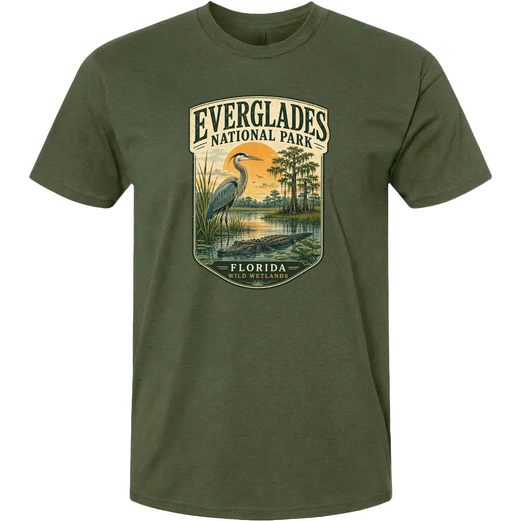 A green t-shirt with a graphic design depicting a bird, a water body, and a landscape with trees and a sunset. The text on the t-shirt reads "EVERGLADES NATIONAL PARK FLORIDA WILD WETLANDS".