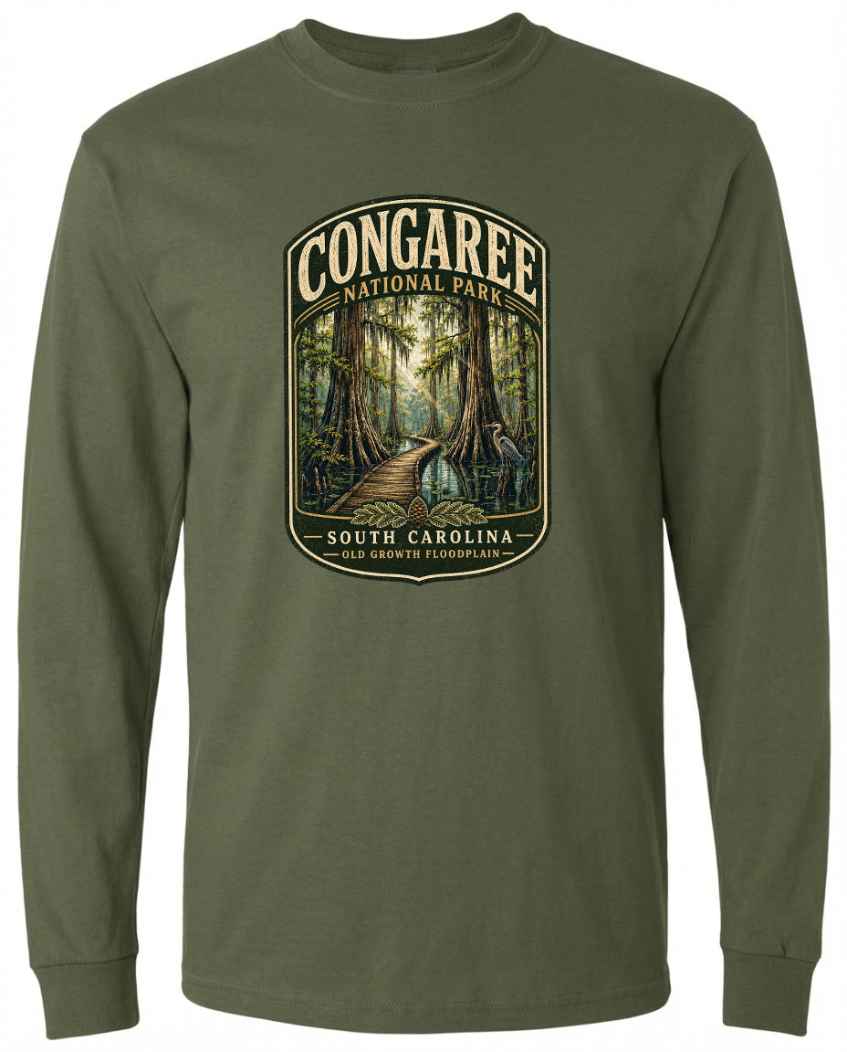 A green long-sleeved t-shirt with a graphic design depicting a forest scene, featuring a path leading through the trees and the text "CONGAREE NATIONAL PARK SOUTH CAROLINA OLD GROWTH FLOODPLAIN".