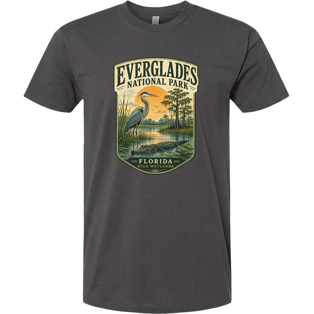 A gray t-shirt with a graphic design depicting a bird, a body of water, and a landscape with trees and a sunset. The text on the t-shirt reads "EVERGLADES NATIONAL PARK FLORIDA WILD WETLANDS".