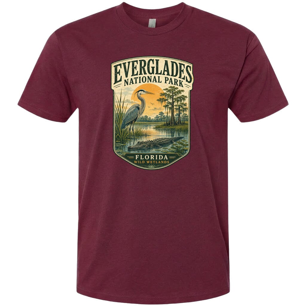 A maroon t-shirt with a graphic design depicting a bird standing in a marshy area with a body of water and trees in the background.