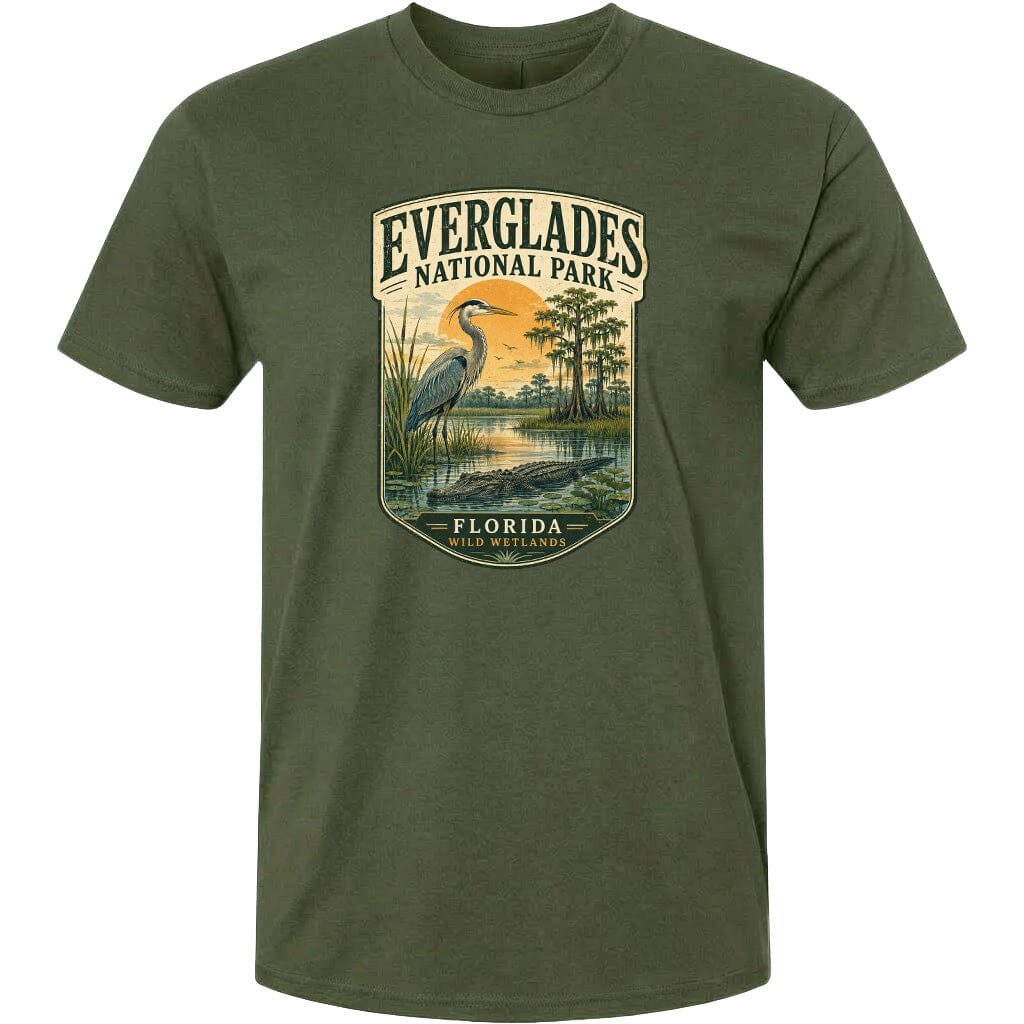 A green t-shirt with a graphic design depicting a bird, a water body, and a landscape with trees and a sunset. The text on the t-shirt reads "EVERGLADES NATIONAL PARK FLORIDA WILD WETLANDS".