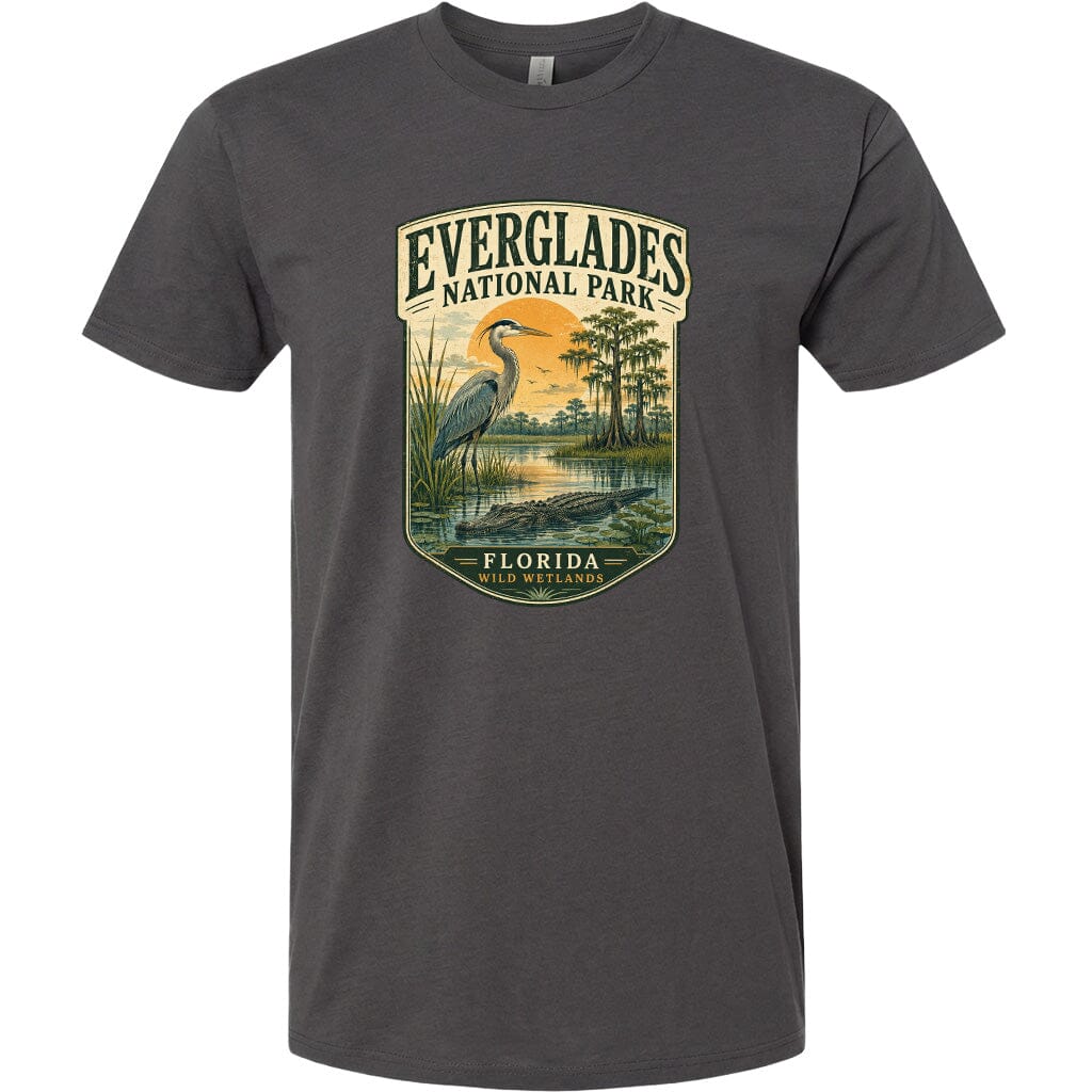 A gray t-shirt with a graphic design depicting a bird, a body of water, and a landscape with trees and a sunset. The text on the t-shirt reads "EVERGLADES NATIONAL PARK FLORIDA WILD WETLANDS".