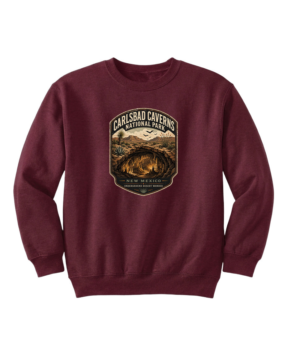A burgundy-colored sweatshirt with a graphic design featuring a shield-shaped emblem that says "CARLSBAD CAVENS NATIONAL PARK" and includes a desert landscape with mountains in the background.