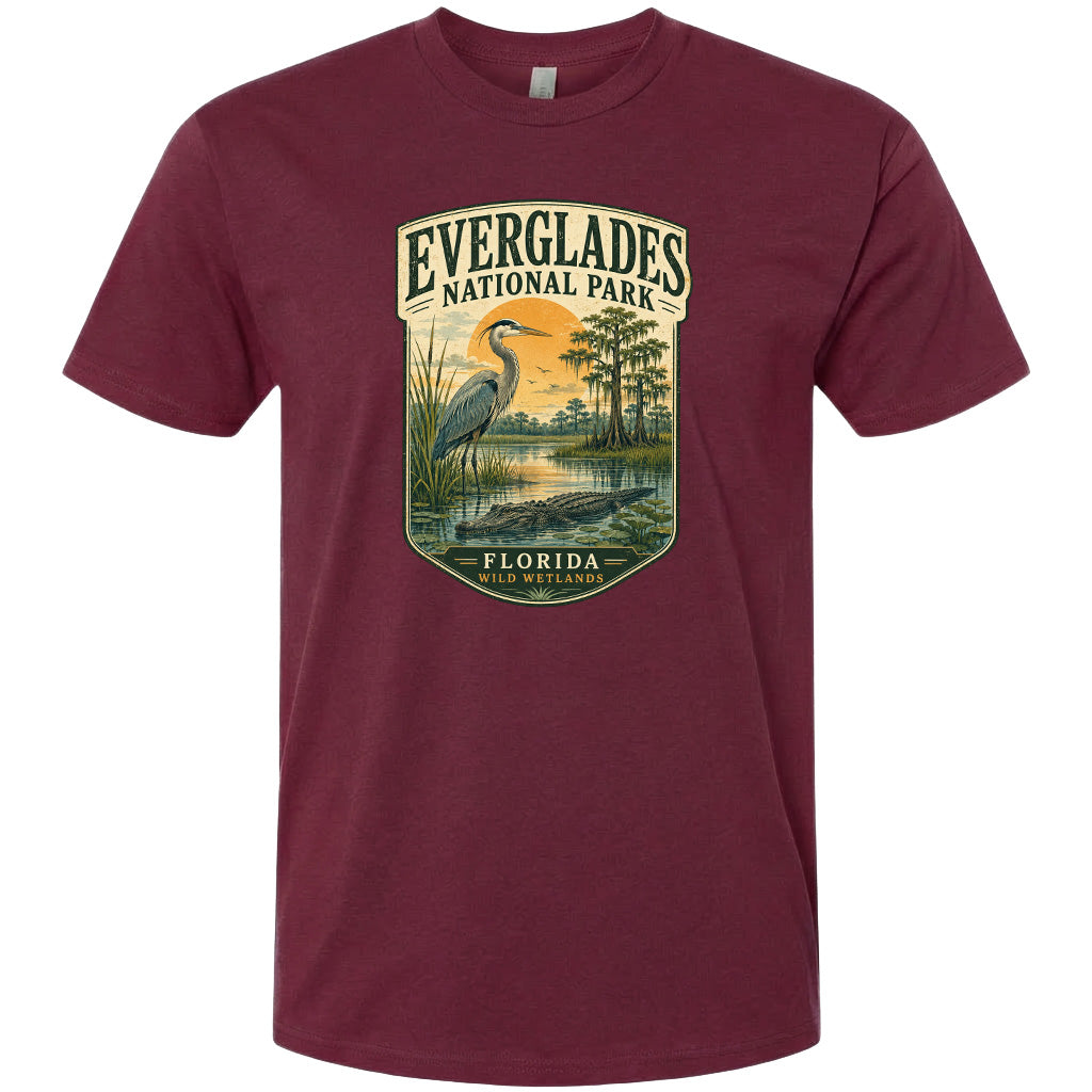 A maroon t-shirt with a graphic design depicting a bird standing in a marshy area with a body of water and trees in the background.