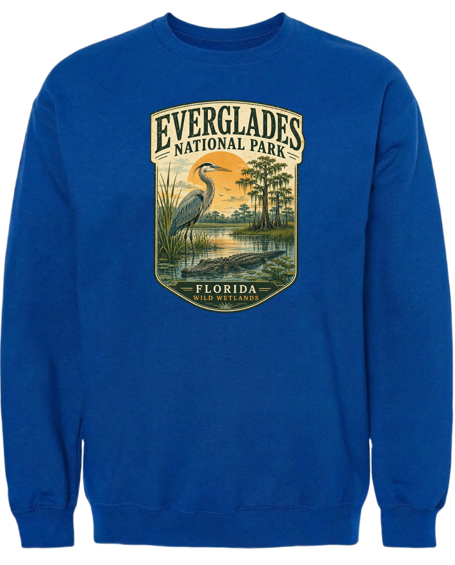 A blue sweatshirt with a graphic design depicting a bird, likely a heron, standing in a marshy area with trees and a sunset in the background.